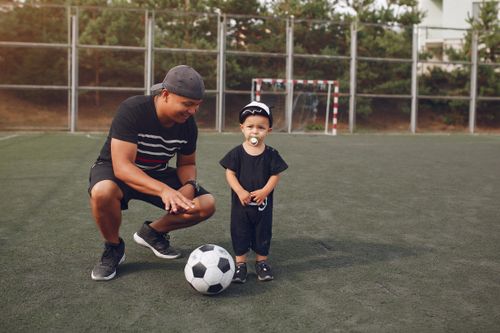 Free Full body of happy ethnic father with football ball hunkering down and teaching son to play soccer while spending time together on football field Stock Photo