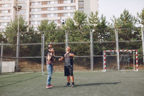 Free Full body cheerful father playing with football ball and mother giving piggyback ride to cute son while walking together on sports ground Stock Photo