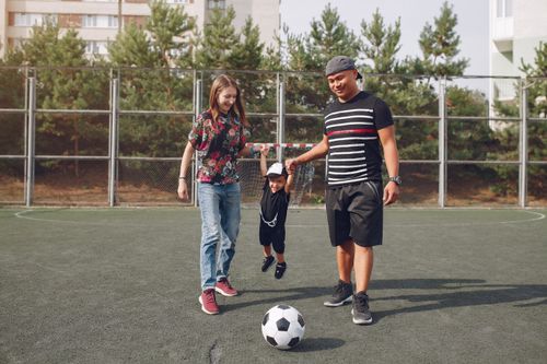 Free Full body of positive content diverse parents holding son hands and teaching boy to kick ball on football field on sunny summer day Stock Photo