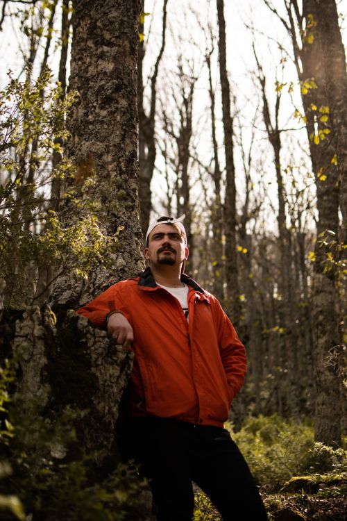Free A man in an orange jacket leans against a tree in a forest during fall. Stock Photo