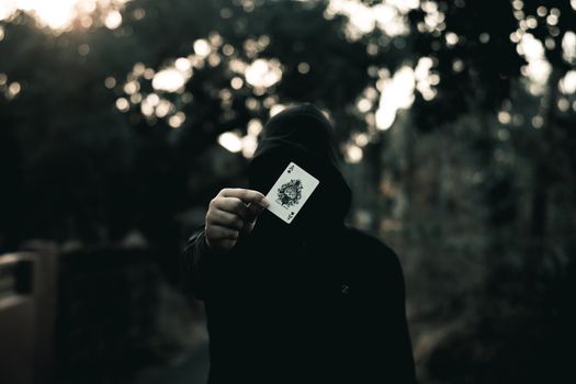 A person in a hoodie holds a playing card, creating a mysterious and dark atmosphere.