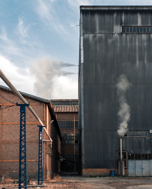 Buildings and storage on territory of industrial factory with smoke emitting from pipes against blue sky