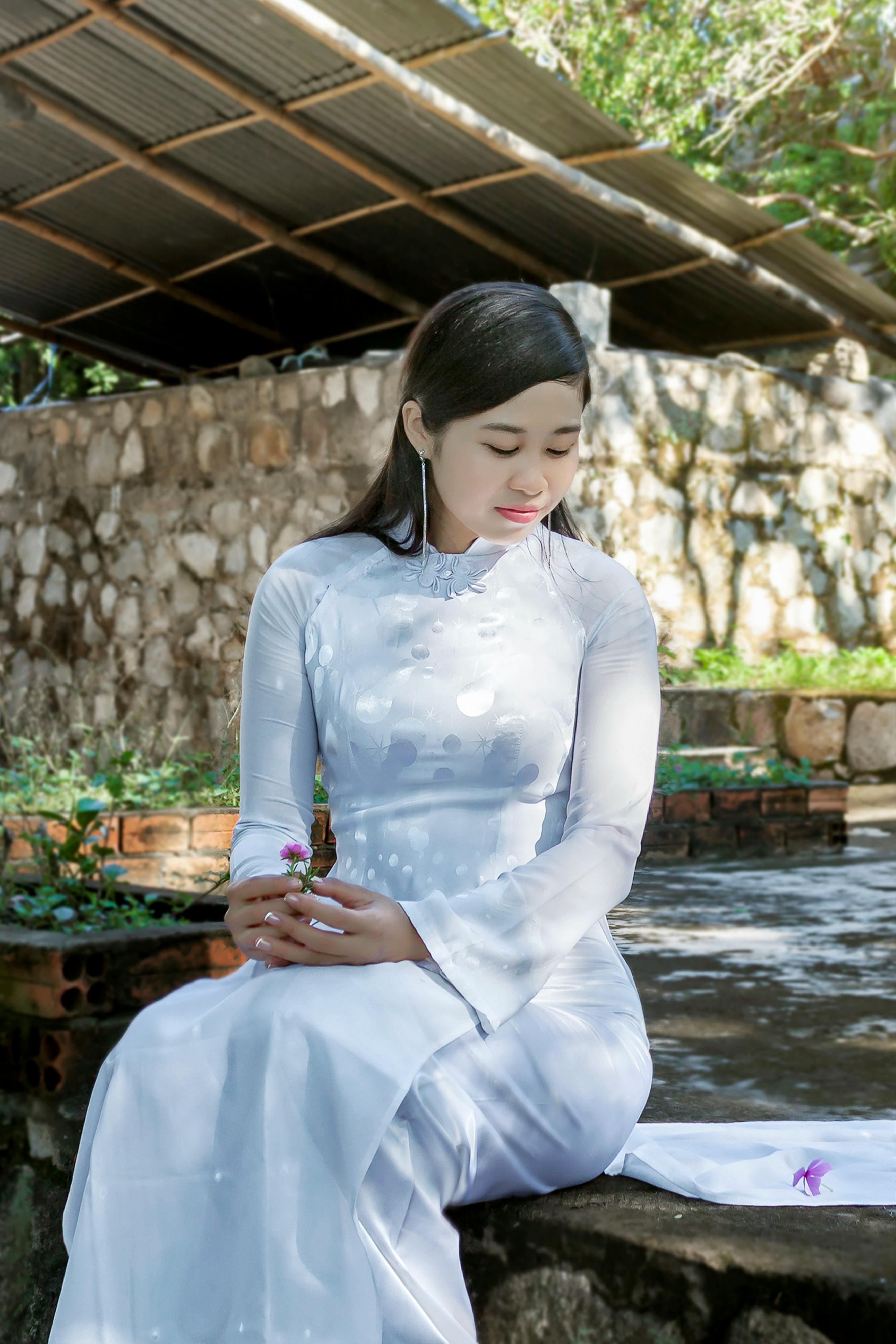 woman in a white long sleeve dress looking down at a flower