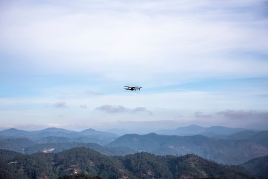 A drone hovering above forested mountains under a clear blue sky.