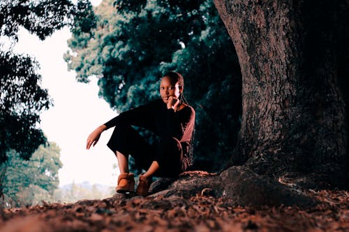 Pensive young African American female in casual clothing looking at camera while sitting under big tree with hand at cheek