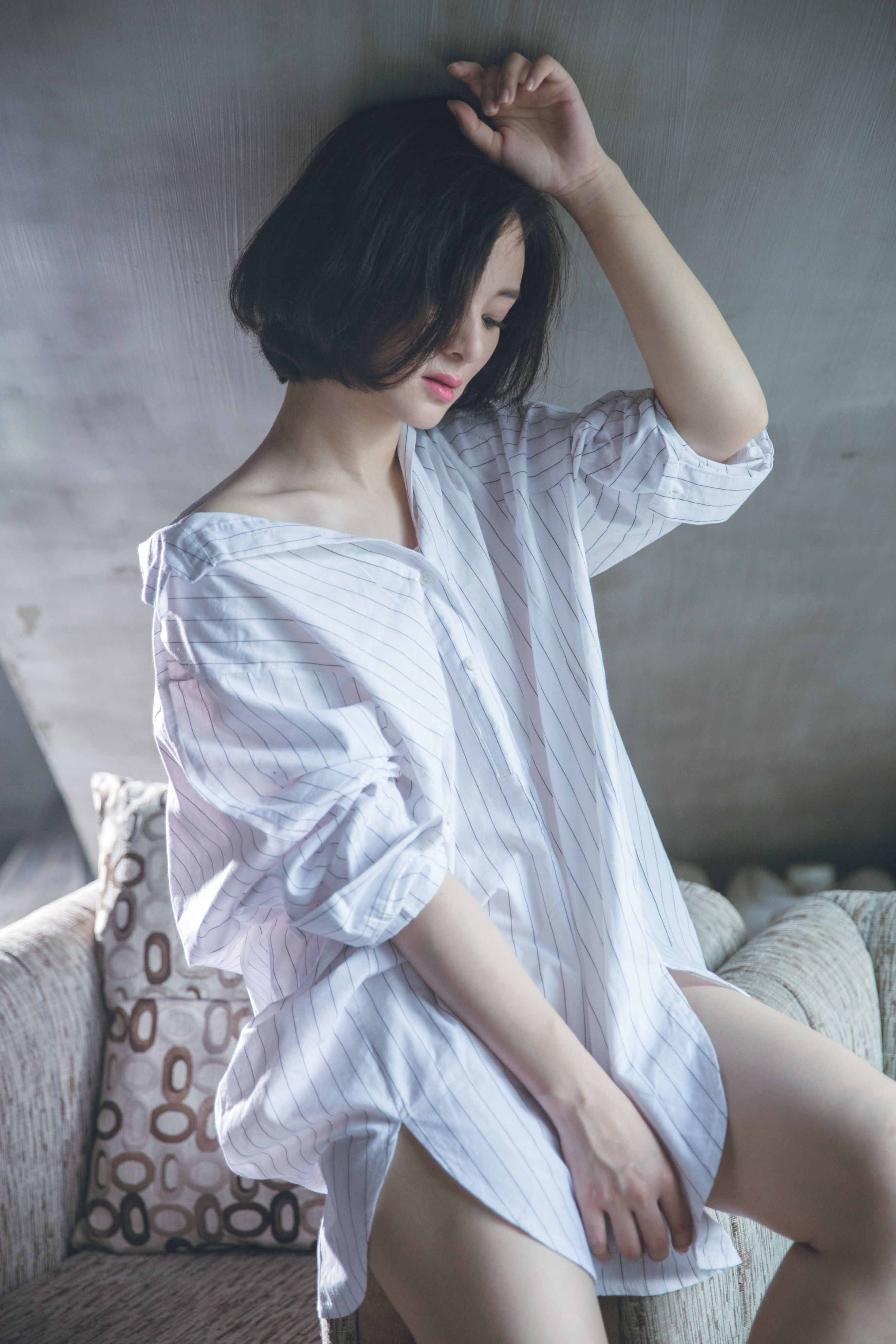 Side view of slender sensitive Asian female in striped shirt looking down while sitting with raised arm on soft armchair edge in house