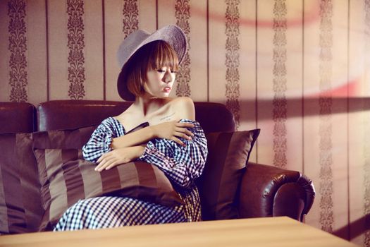 Young slim dreamy female in hat and stylish dress with makeup sitting on comfortable couch with cushion while looking away near decorative wall and wooden table
