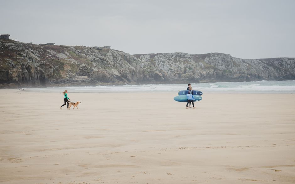 A dog walks on a Brittany beach as a surfer carries surfboards. Scenic and serene