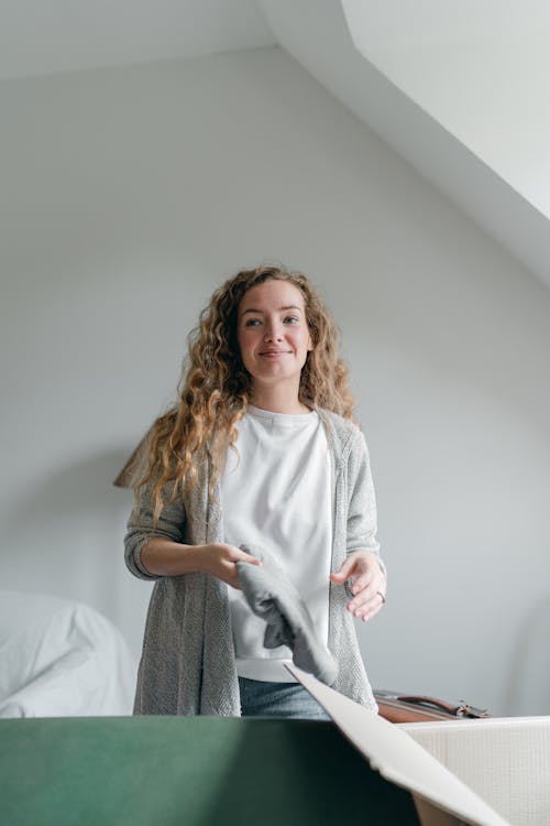 Positive smiling young lady in casual outfit with folded clothes packing stuff into box and suitcase and looking away happily in sunny empty bedroom
