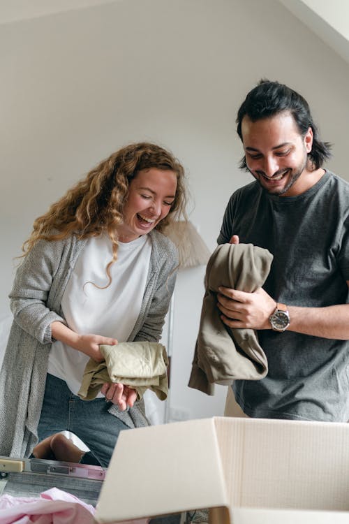 Excited couple in casual clothes packing belongings into carton box and happily laughing while preparing to move into new accommodation together