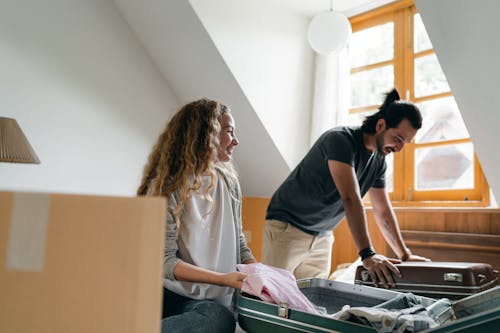 Positive young couple smiling and packing suitcases together in empty bedroom while preparing to move into new house