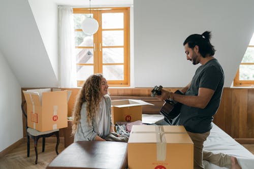 Side view of young bearded content husband playing guitar for smiling wife in bedroom while resting after packing stuff into carton boxes before relocation
