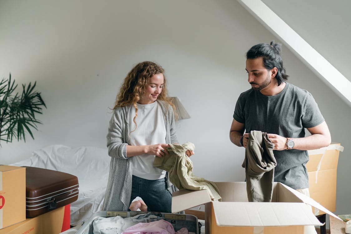Content young couple in casual outfit standing in new modern bedroom and unpacking boxes together after relocation into new apartment