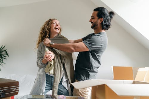 Delighted excited couple in casual outfits having fun and laughing while unpacking stuff in new cozy apartment in daylight
