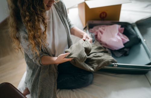 Free From above crop anonymous female in casual wear sitting on bed and packing luggage while preparing to move out Stock Photo