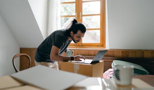 Free Side view of focused bearded male in casual outfit with cup of hot drink browsing laptop while packing stuff into carton boxes Stock Photo