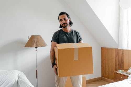 Cheerful young bearded male in casual wear carrying cardboard container in sunny bedroom while moving into new apartment