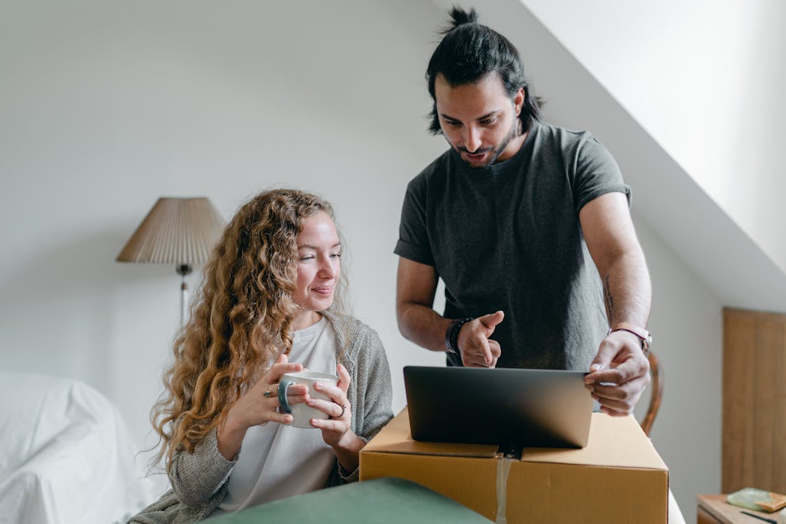 Free Positive happy young couple in casual clothes packing belongings into carton box and using laptop in living room near window Stock Photo