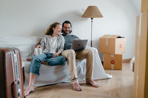 Free Cheerful couple watching video on laptop together while sitting on couch in living room when moving house Stock Photo