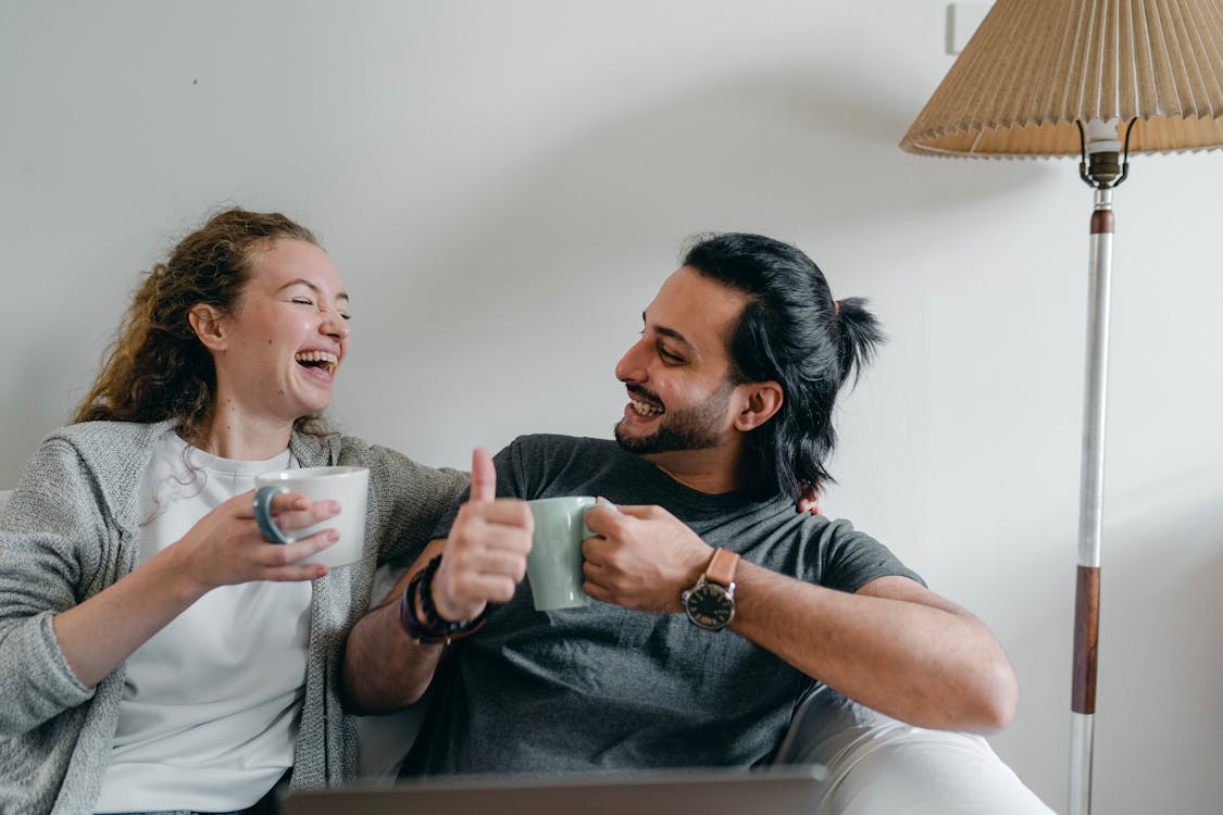 Free Positive girlfriend in casual wear looking at content ethnic man in smart watch while sitting on cozy sofa with cups of hot beverage and looking at each other Stock Photo