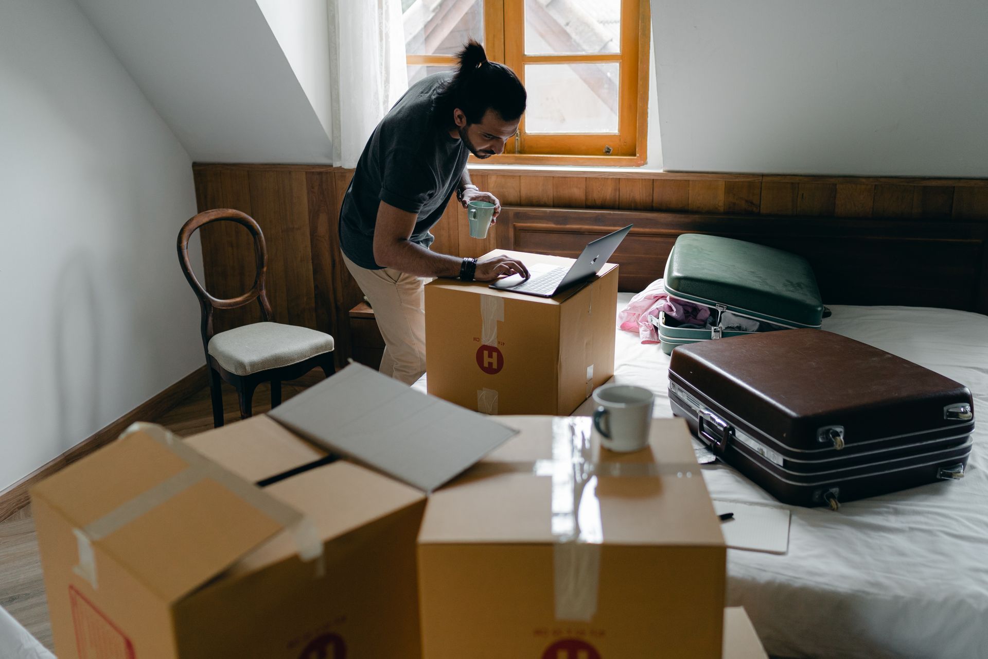 Side view of ethnic male in casual clothes and wristwatch surfing internet on netbook and standing with cup of hot drink surrounded by cardboard boxes and suitcases in bedroom
