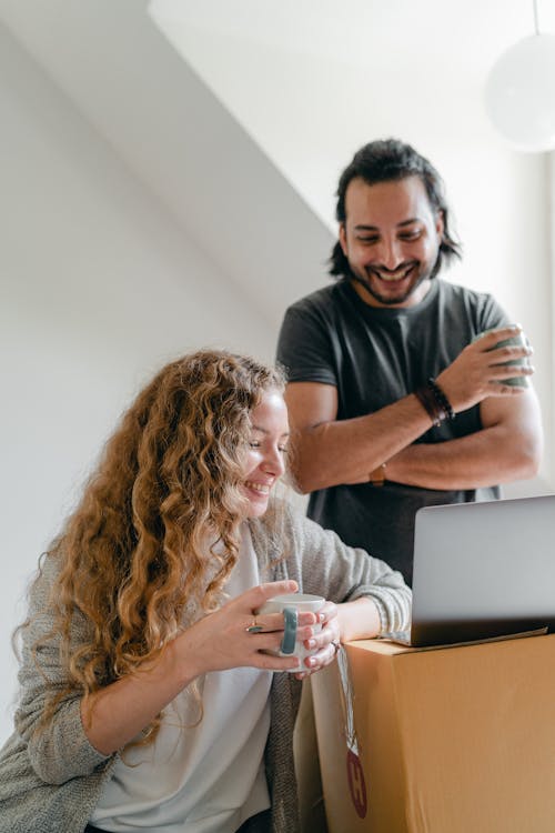 Happy ethnic man in smart watch standing near cheerful girlfriend in casual clothes while watching portable computer and drinking hot beverage in light room in apartment