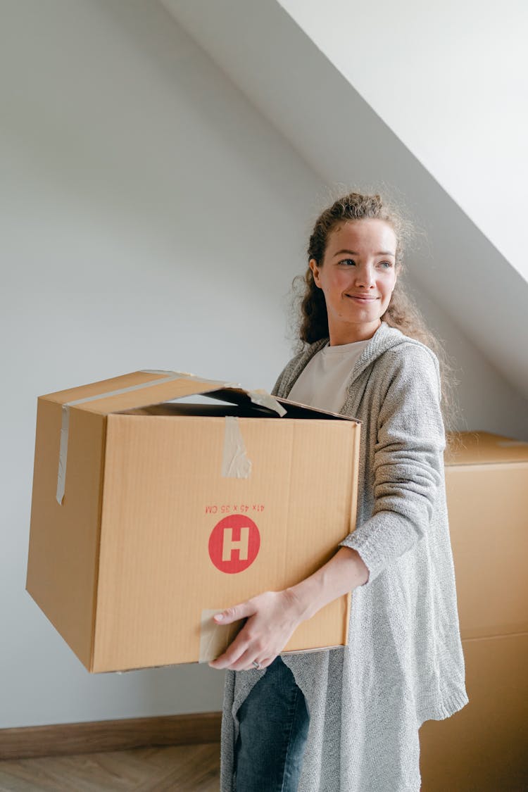 Smiling Woman In Cardigan Holding Cardboard Box At Home