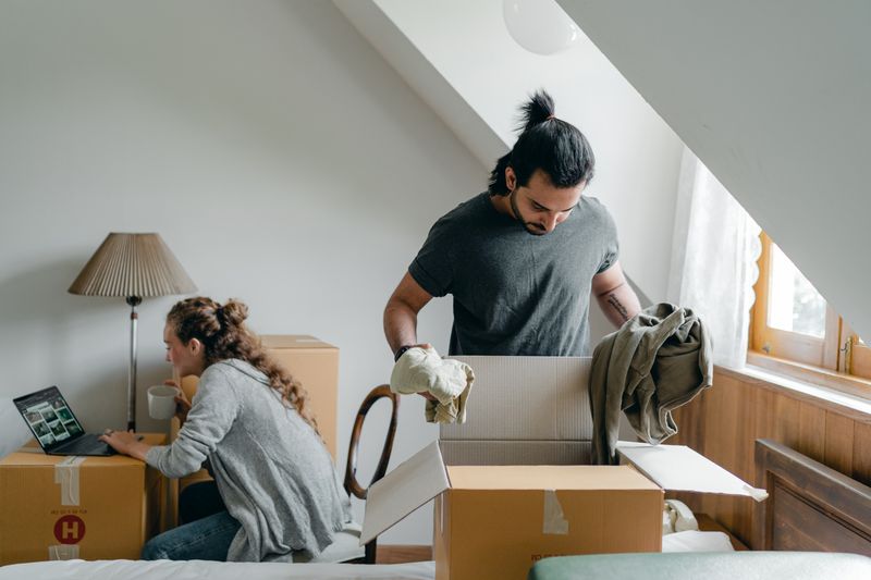 Ethnic male getting clothes out of cardboard box and girlfriend watching photo gallery on netbook while drinking coffee in attic style room