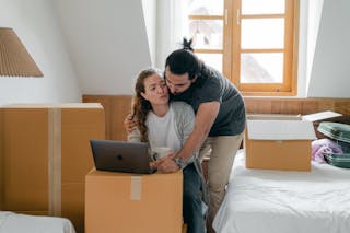 Ethnic boyfriend in wristwatch cuddling girlfriend while watching netbook and standing near cardboard boxes and bed with suitcase after moving into new house