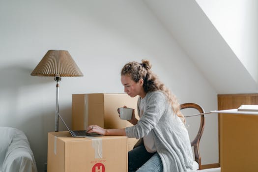 Side view of thoughtful female in cardigan browsing internet on portable computer while sitting on chair near big cardboard boxes and floor lamp in light flat