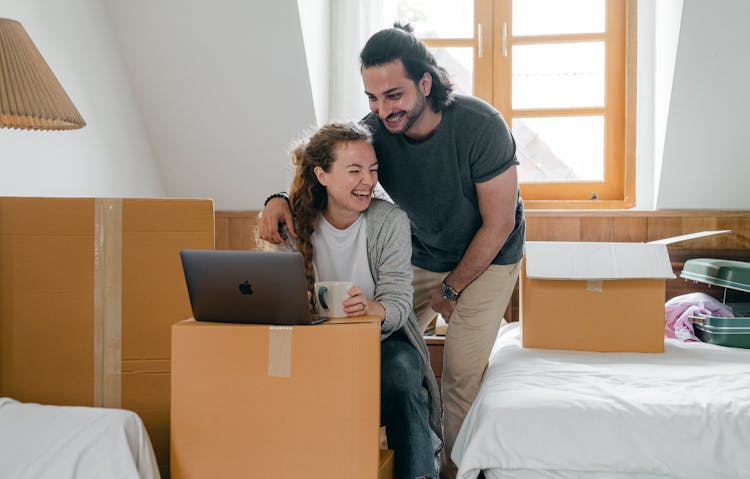 Happy Diverse Couple With Laptop Sitting In Attic Style Bedroom