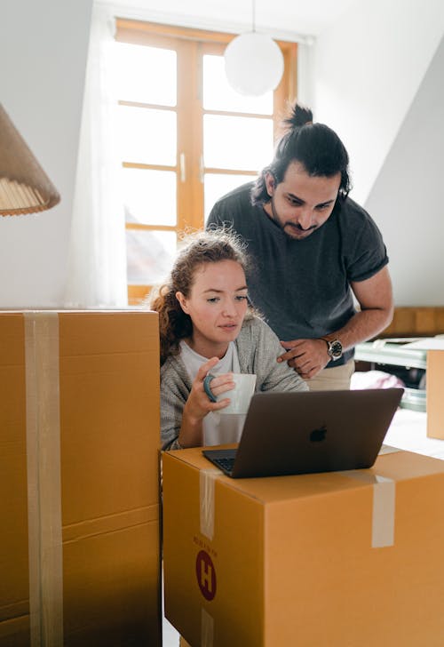 Free Thoughtful woman surfing internet on netbook while sitting with cup of hot drink near ethnic boyfriend in watch and casual wear standing near cardboard boxes after moving into new flat Stock Photo