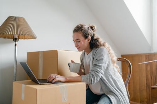 Content lady in casual wear typing on netbook while sitting on chair with cup of hot drink near big cardboard boxes in light apartment