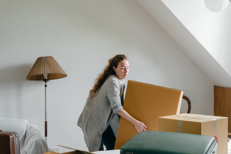 Woman Lifting Heavy Cardboard Box While Standing Near Floor Lamp