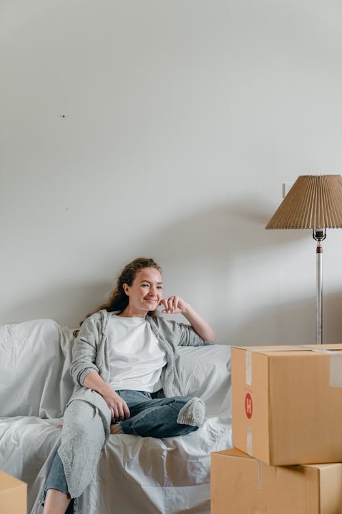 Free Happy female in jeans and cardigan sitting with crossed legs leaned on hand on comfortable couch in front of pile of cardboard boxed near white wall and looking away Stock Photo