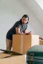 Thoughtful ethnic male in casual clothes standing leaned on big square cardboard box near suitcase and looking at camera in apartment