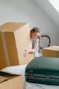 Female in casual clothes and ring on finger peeping out of big cardboard box while standing leaned forward near bed with suitcase in attic style room