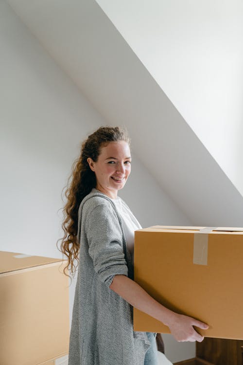 Side view of cheerful female in cardigan with wavy hair holding big cardboard box and looking at camera in attic style living room