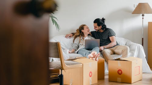 Free Cheerful couple in casual clothes sitting on comfortable sofa with laptop and looking at each other while moving to new apartment Stock Photo