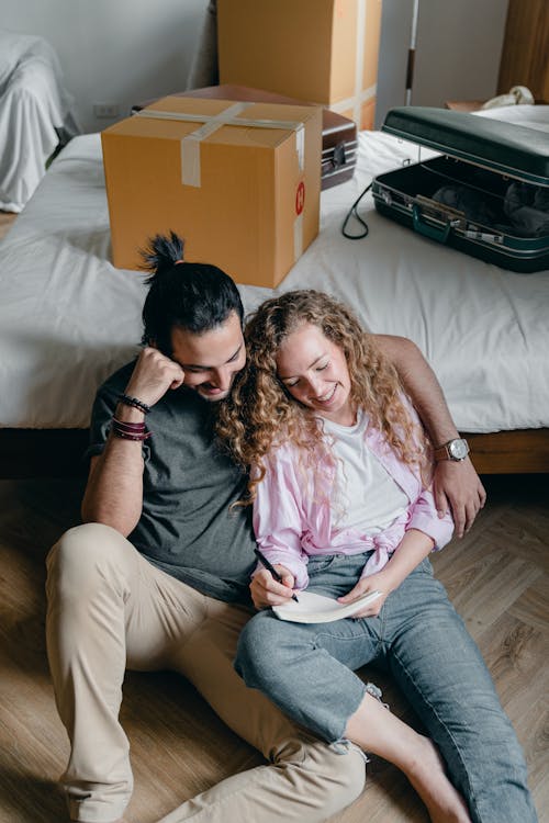 Free From above of candid couple sitting on floor near bed and writing in notebook while packing things Stock Photo