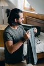 Smiling bearded ethnic male in casual t shirt taking out jeans from opened carton box while unpacking clothes in attic bedroom of new apartment after moving and looking away