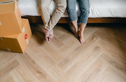 Unrecognizable barefoot man and woman sitting on bed near carton boxes while relaxing from unboxing belongings during relocation in new flat