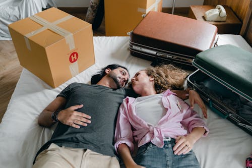 Free From above of young ethnic bearded man and woman with curly hair looking at each other and lying on bed among suitcases and cardboard boxes with stuff while moving in new apartment together Stock Photo