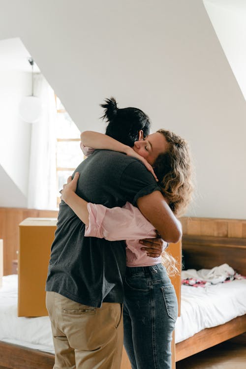 Loving young couple embracing while standing in cozy attic bedroom near bed with unpacked cardboard box while moving in new house