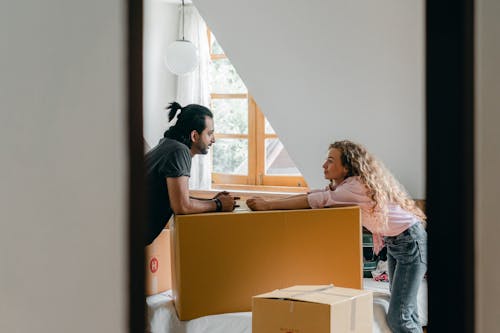 Side view from entrance of cheerful young ethnic bearded man with ponytail and woman with curly hair leaning on large cardboard package while arranging stuff in cozy attic bedroom