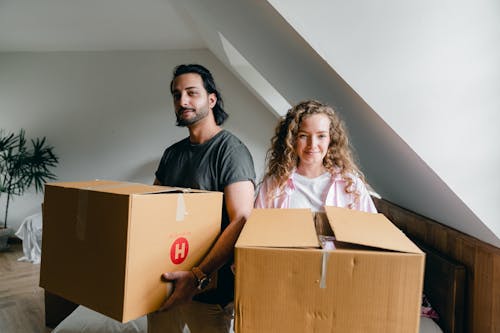 Young woman and ethnic man in casual clothes smiling at camera and holding big boxes during relocation to new apartment