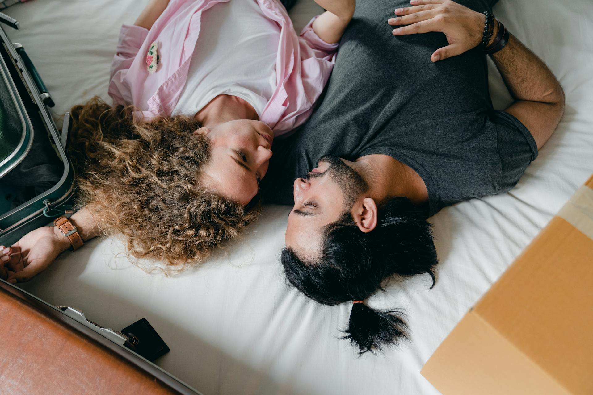 From above of multiethnic couple in casual clothes lying near suitcases on bed while resting and hugging during moving into new apartment