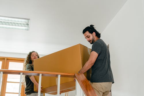 From below of couple in casual clothes carrying big carton box together while moving packed personal items into new apartment