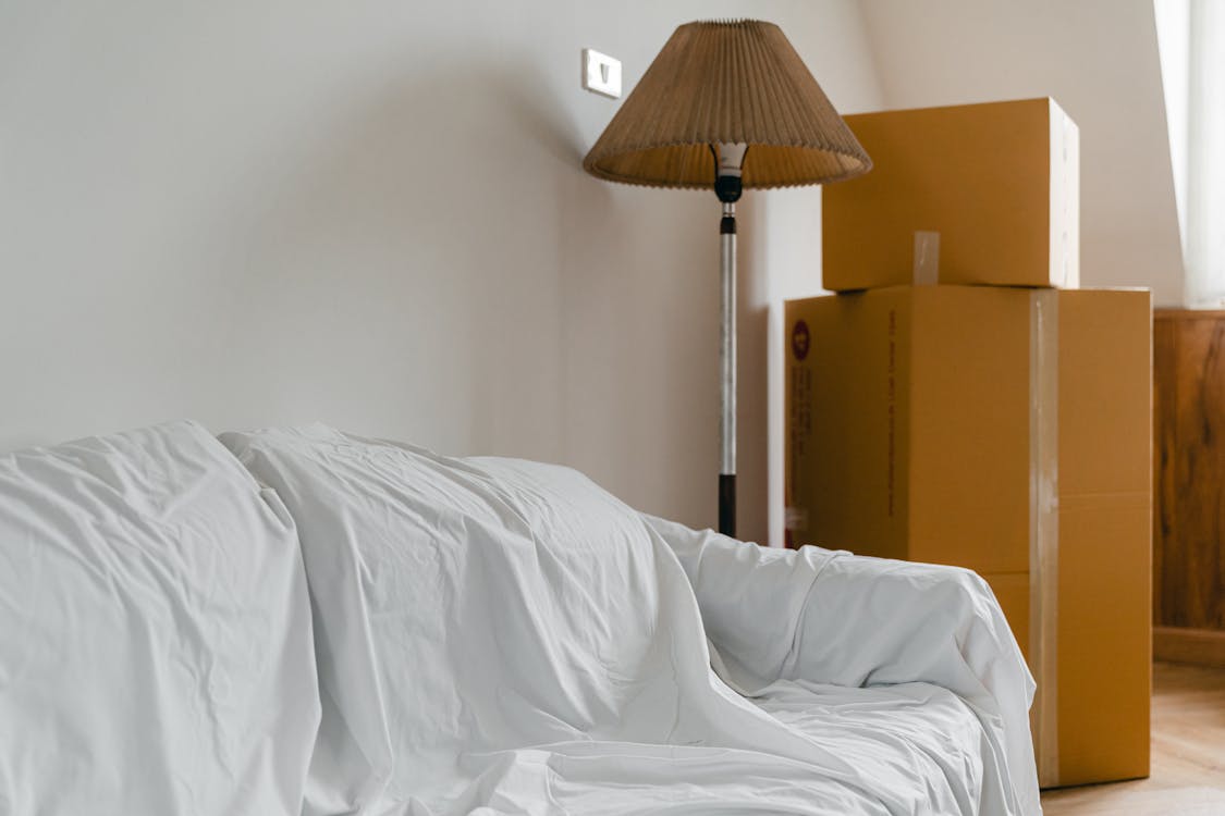 Covered with sheet sofa and floor lamp and packed cardboard boxes in light spacious room of new contemporary house