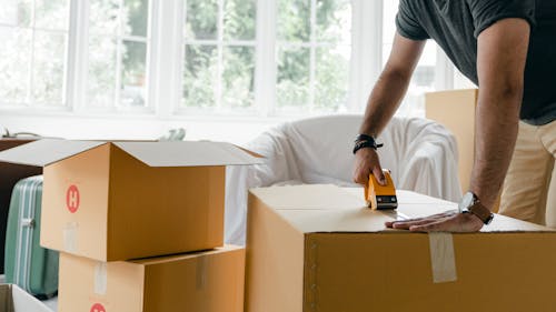 Crop unrecognizable man in casual clothes packing carton box using scotch tape dispenser for moving personal items to new apartment